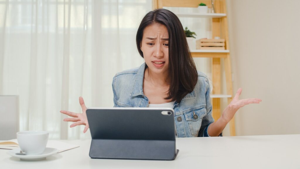 Frustrated Young Asia lady having problem with not working tablet computer sitting on desk.